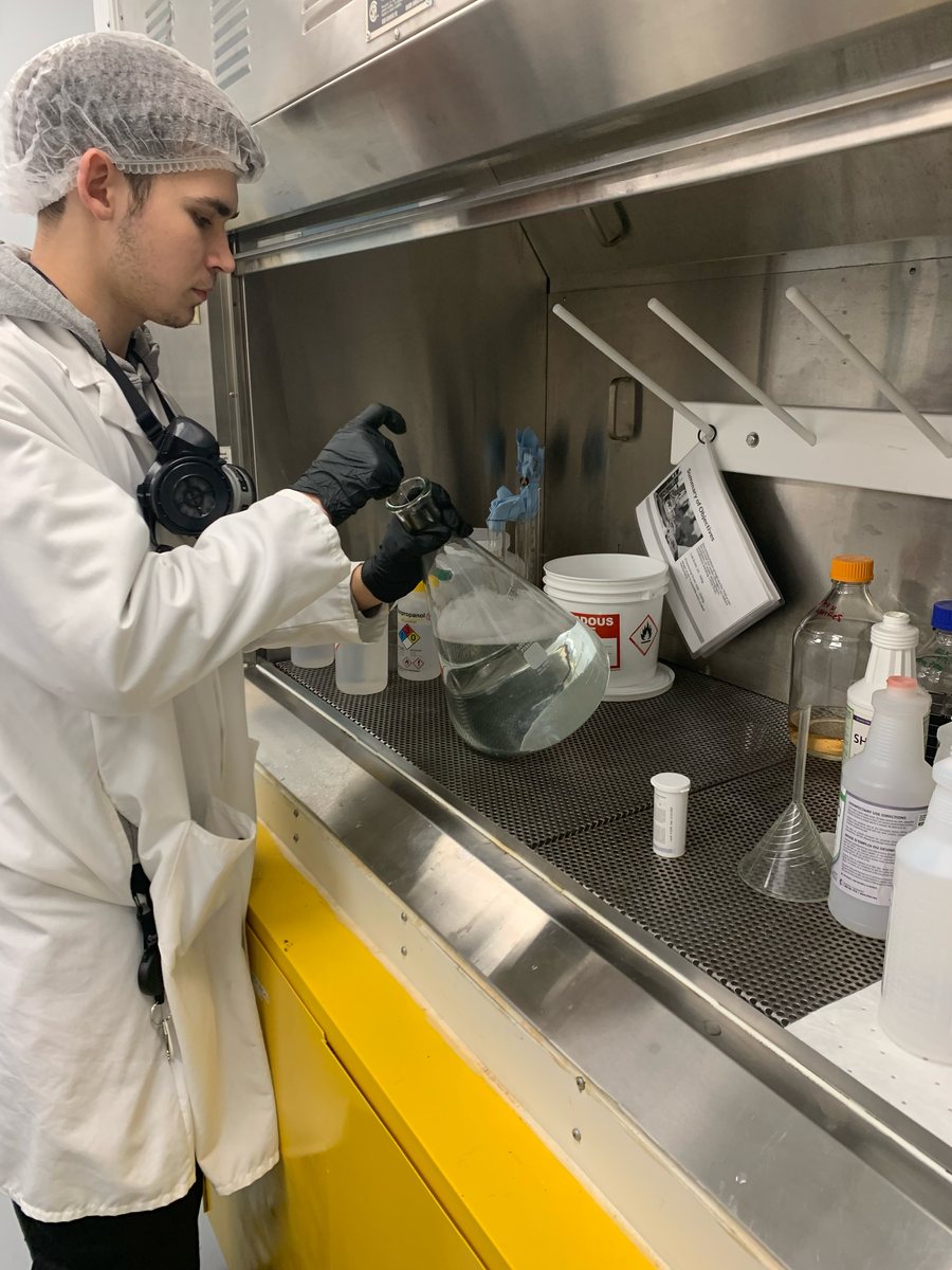 A technician in a hairnet, lab coat and gloves working at a stainless steel fume hood with glassware and labelled solvent bottles
