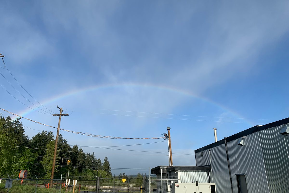A rainbow arcing over the Protonify facility, a grey metal building beside a fenced yard and treeline